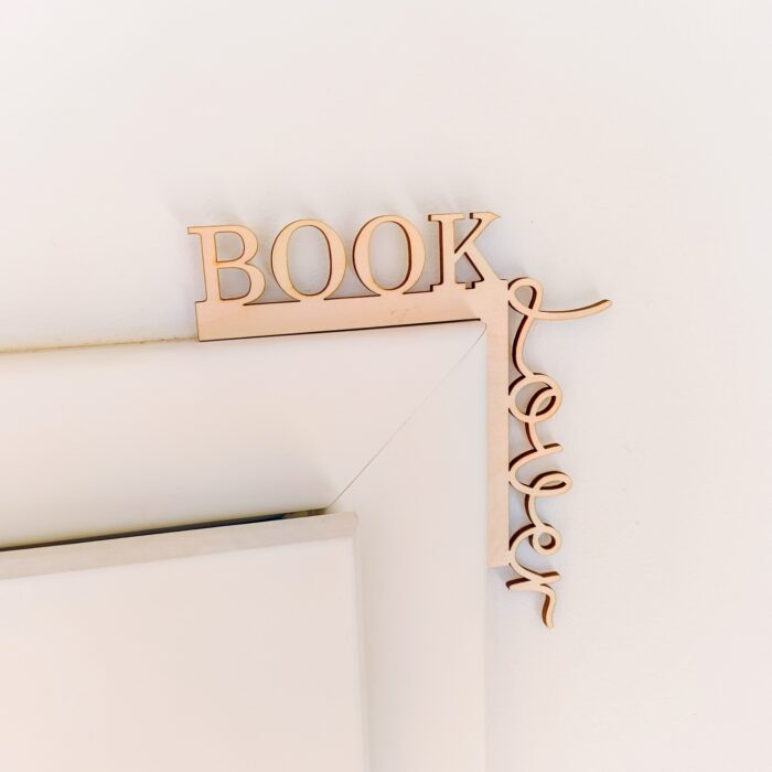 Handgefertigte Türrahmen-Deko aus hellem Holz mit dem Schriftzug „Book Lover“ im Detail. Der Aufsatz sitzt auf der oberen linken Ecke eines weißen Holzrahmens vor einer strukturierten, weißen Wand. Das Wort „BOOK“ verläuft vertikal nach unten, während „Lover“ in geschwungener Schreibschrift horizontal auf dem Rahmen aufliegt.