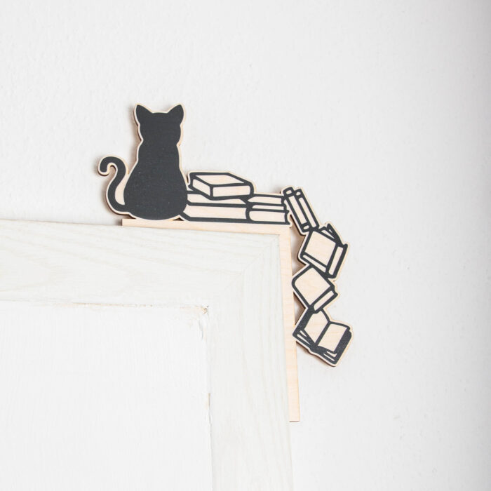 Schwarze Silhouette einer sitzenden Katze auf einem Stapel Bücher als dekorativer Aufsatz für die rechte obere Ecke eines weißen Tür- oder Bilderrahmens. Die Deko besteht aus hellem Holz mit schwarzem Aufdruck, wobei einige Bücher scheinbar die Ecke hinunterfallen.