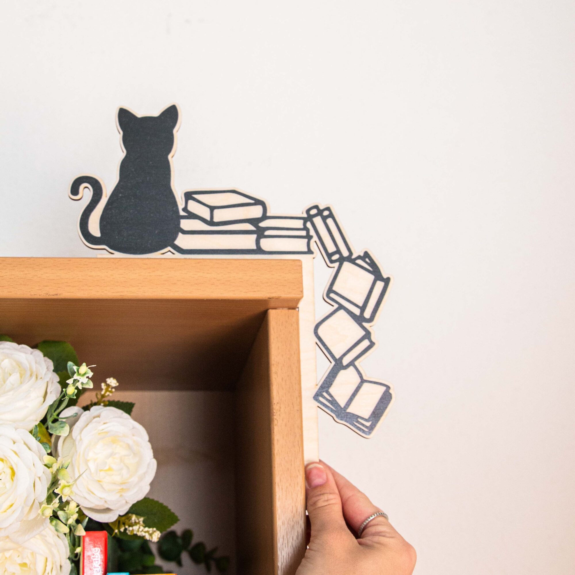 Türrahmen Deko Katze und Bücher | Türaufsatz Holz Silhouette | Geschenk für Katzenliebhaber & Leseratten, Regal Deko 5 Schwarze Silhouette einer sitzenden Katze auf einem Stapel Bücher als dekorativer Aufsatz für die rechte obere Ecke eines weißen Tür- oder Bilderrahmens. Die Deko besteht aus hellem Holz mit schwarzem Aufdruck, wobei einige Bücher scheinbar die Ecke hinunterfallen.