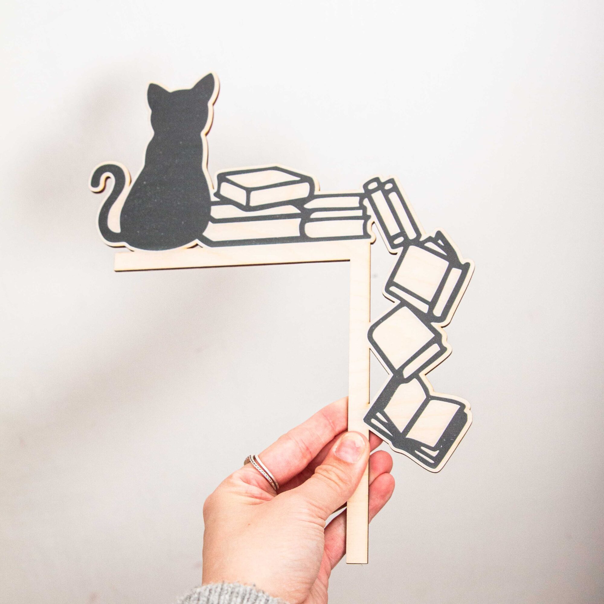 Türrahmen Deko Katze und Bücher | Türaufsatz Holz Silhouette | Geschenk für Katzenliebhaber & Leseratten, Regal Deko 3 Schwarze Silhouette einer sitzenden Katze auf einem Stapel Bücher als dekorativer Aufsatz für die rechte obere Ecke eines weißen Tür- oder Bilderrahmens. Die Deko besteht aus hellem Holz mit schwarzem Aufdruck, wobei einige Bücher scheinbar die Ecke hinunterfallen.