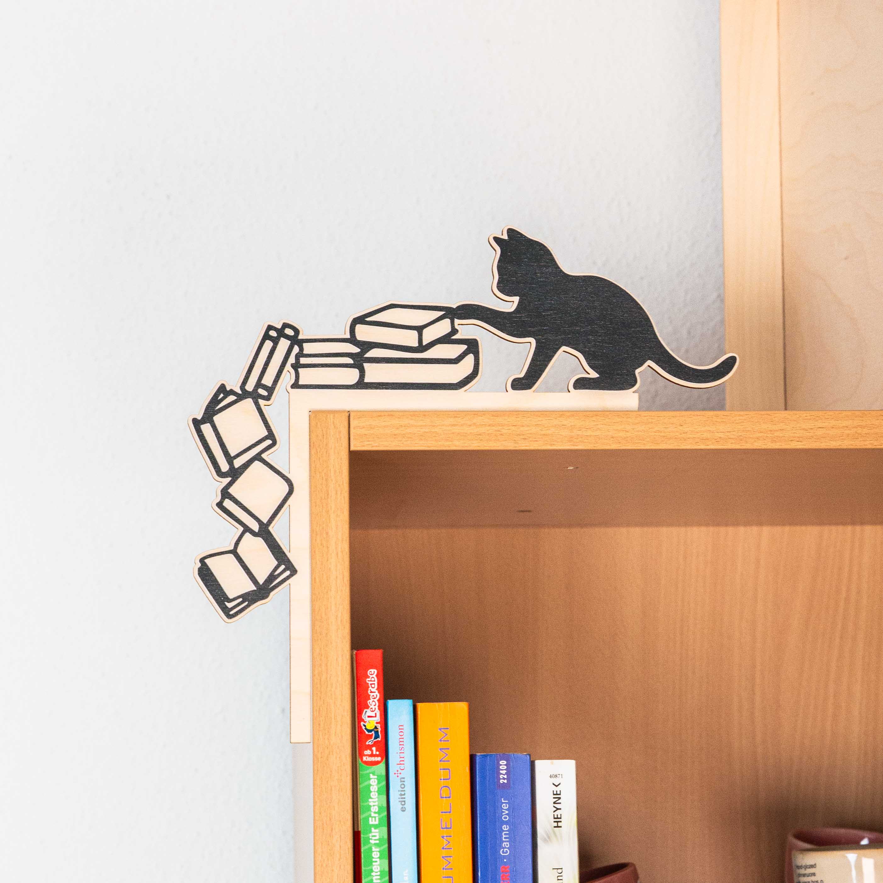 Regal- & Türrahmen-Deko „Freche Katze“ 6 Handgefertigter Regal-Aufsatz aus Holz an der oberen linken Ecke eines hellen Holzregals. Das Design zeigt die schwarze Silhouette einer Katze, die mit der Pfote einen Stapel Bücher anstößt. Links daneben stürzen vier gezeichnete Bücher in einer dynamischen Kurve die Seitenkante des Regals hinunter. Eine Hand hält das Produkt am Rahmen fest, im Regal darunter stehen bunte Buchrücken.