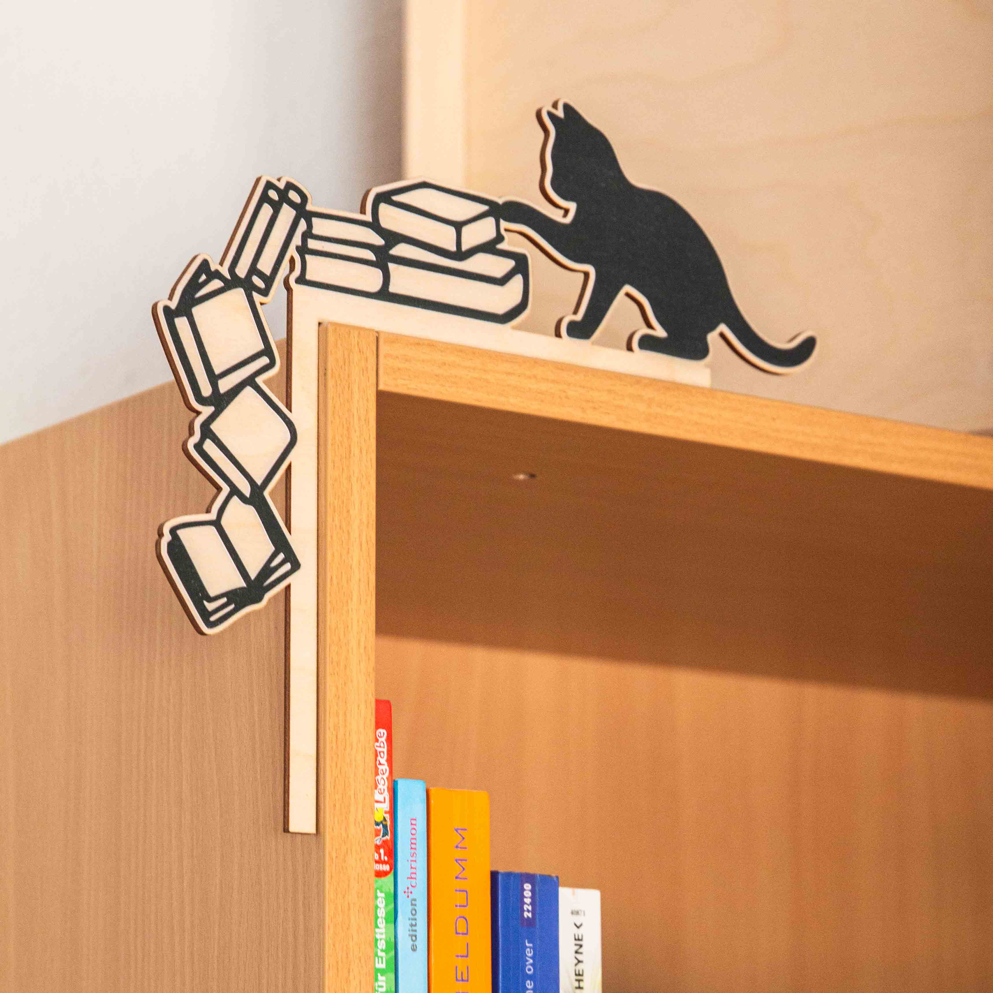 Regal- & Türrahmen-Deko „Freche Katze“ 5 Handgefertigter Regal-Aufsatz aus Holz an der oberen linken Ecke eines hellen Holzregals. Das Design zeigt die schwarze Silhouette einer Katze, die mit der Pfote einen Stapel Bücher anstößt. Links daneben stürzen vier gezeichnete Bücher in einer dynamischen Kurve die Seitenkante des Regals hinunter. Eine Hand hält das Produkt am Rahmen fest, im Regal darunter stehen bunte Buchrücken.