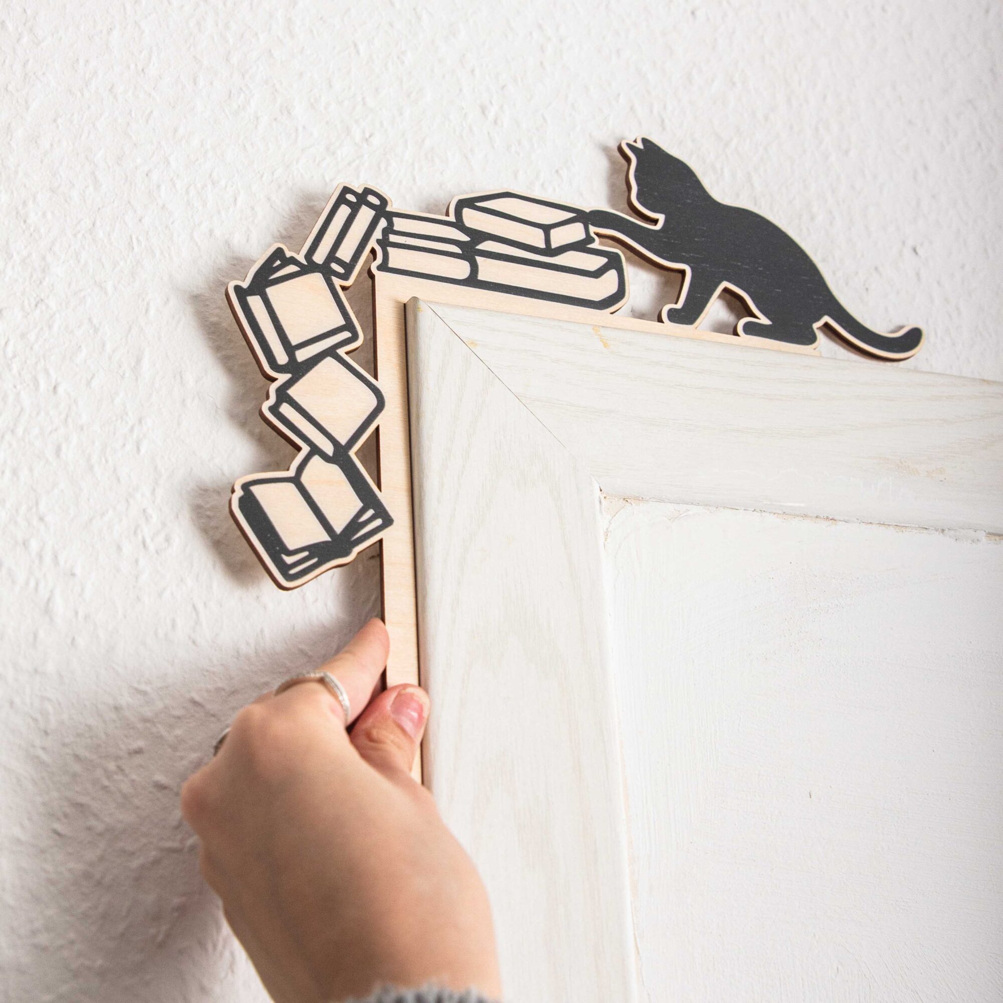 Regal- & Türrahmen-Deko „Freche Katze“ 4 Handgefertigter Regal-Aufsatz aus Holz an der oberen linken Ecke eines hellen Holzregals. Das Design zeigt die schwarze Silhouette einer Katze, die mit der Pfote einen Stapel Bücher anstößt. Links daneben stürzen vier gezeichnete Bücher in einer dynamischen Kurve die Seitenkante des Regals hinunter. Eine Hand hält das Produkt am Rahmen fest, im Regal darunter stehen bunte Buchrücken.