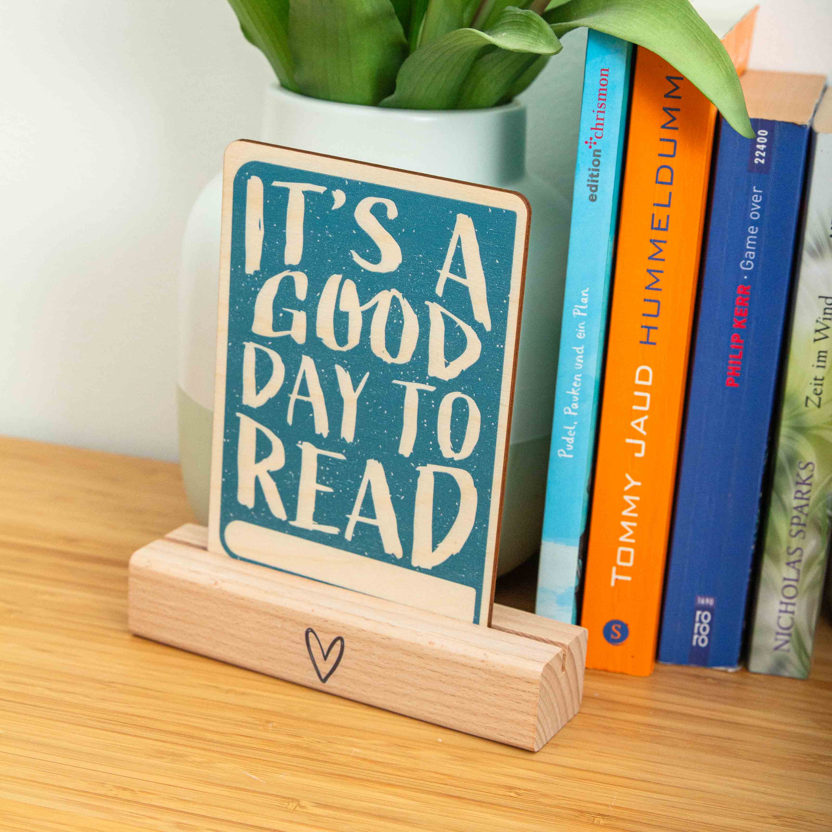 Postkarte Its a good day to read / Holzpostkarte Bücherliebhaber / Bookstagram / Postkarten mit Botschaft 1 Eine hochkant ausgerichtete Holzpostkarte liegt flach auf einer hellbraunen Holzoberfläche mit horizontaler Maserung. Die Karte hat einen vollflächigen, türkisblauen Hintergrund mit einer leichten Textur. Der englische Spruch „IT'S A GOOD DAY TO READ“ ist in großen, unregelmäßigen Holzbuchstaben (Negativdruck) ausgespart. Am unteren Rand befindet sich ein schmaler, horizontaler Balken in der natürlichen Holzfarbe, der Platz für eine Beschriftung lässt.