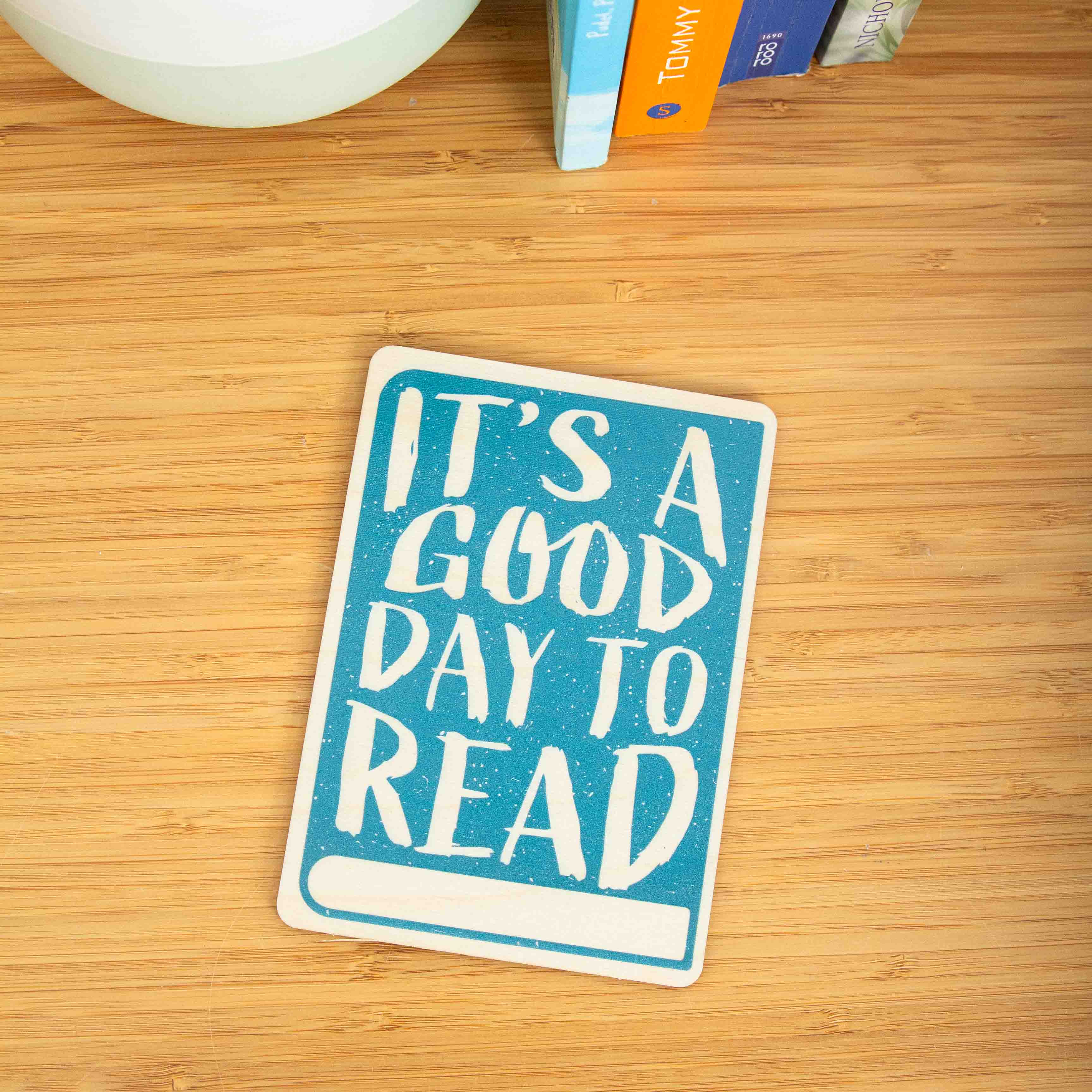 Postkarte Its a good day to read / Holzpostkarte Bücherliebhaber / Bookstagram / Postkarten mit Botschaft 3 Eine hochkant ausgerichtete Holzpostkarte liegt flach auf einer hellbraunen Holzoberfläche mit horizontaler Maserung. Die Karte hat einen vollflächigen, türkisblauen Hintergrund mit einer leichten Textur. Der englische Spruch „IT'S A GOOD DAY TO READ“ ist in großen, unregelmäßigen Holzbuchstaben (Negativdruck) ausgespart. Am unteren Rand befindet sich ein schmaler, horizontaler Balken in der natürlichen Holzfarbe, der Platz für eine Beschriftung lässt.