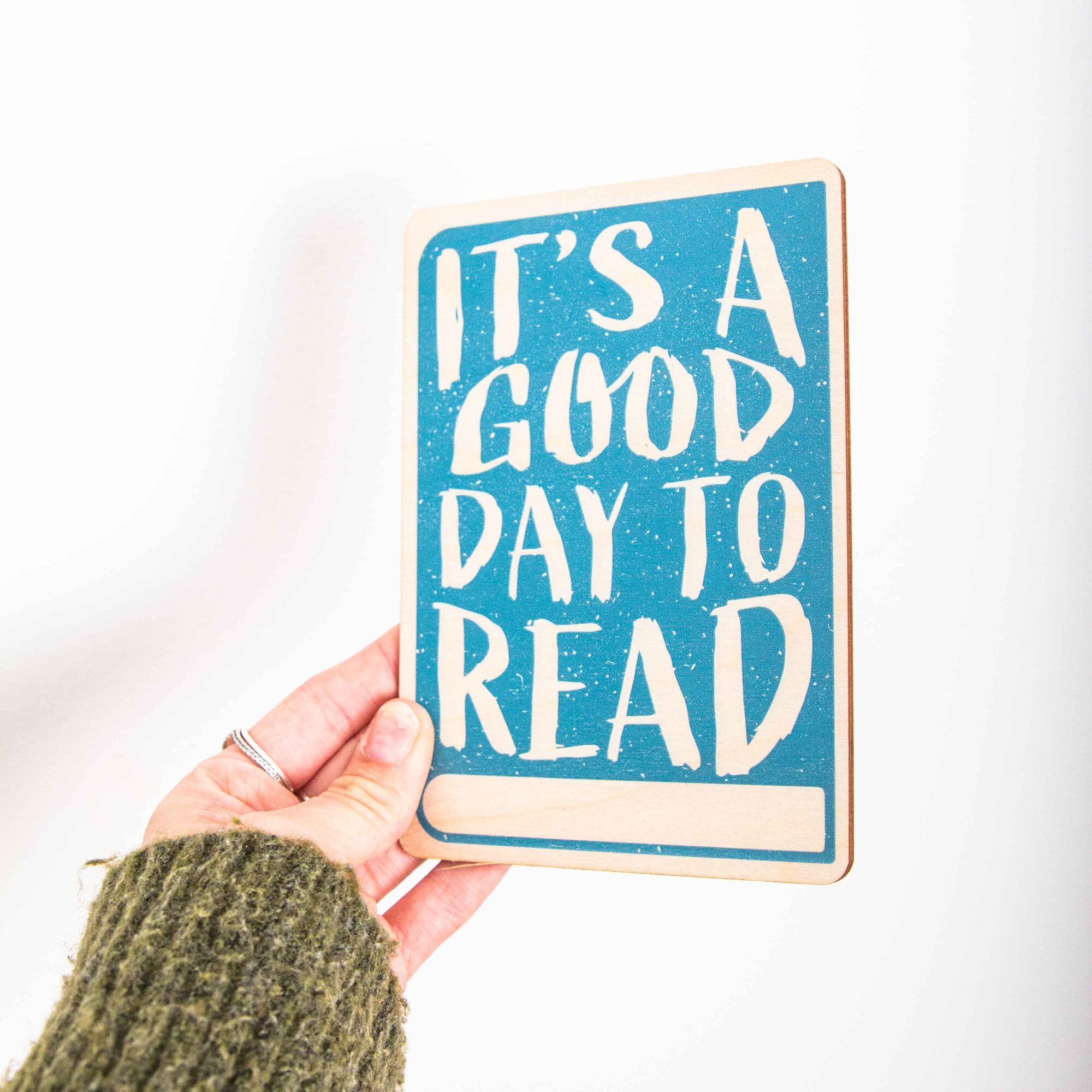 Postkarte Its a good day to read / Holzpostkarte Bücherliebhaber / Bookstagram / Postkarten mit Botschaft 2 Eine hochkant ausgerichtete Holzpostkarte liegt flach auf einer hellbraunen Holzoberfläche mit horizontaler Maserung. Die Karte hat einen vollflächigen, türkisblauen Hintergrund mit einer leichten Textur. Der englische Spruch „IT'S A GOOD DAY TO READ“ ist in großen, unregelmäßigen Holzbuchstaben (Negativdruck) ausgespart. Am unteren Rand befindet sich ein schmaler, horizontaler Balken in der natürlichen Holzfarbe, der Platz für eine Beschriftung lässt.