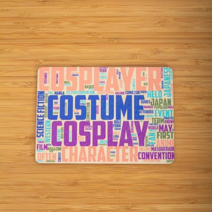 Eine rechteckige Holzpostkarte im Querformat. Im Zentrum stechen vier Begriffe in sehr großen, strukturierten Großbuchstaben hervor: „COSPLAYER“ in Lachsrosa ganz oben, „COSTUME“ in Dunkelblau darunter, gefolgt von „COSPLAY“ in Lila und „CHARACTER“ in hellem Orange am unteren Rand. Die Zwischenräume und Ränder sind dicht mit kleineren, zugehörigen englischen Begriffen wie „Manga“, „Anime“, „Convention“, „Japan“ und „Science Fiction“ in verschiedenen Farben und Ausrichtungen gefüllt.