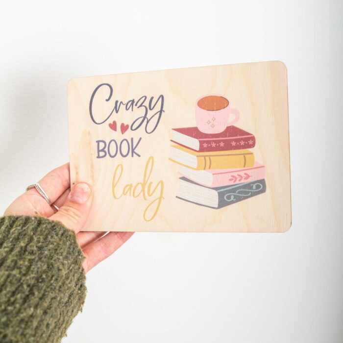 Eine rechteckige Holzpostkarte im Querformat liegt flach auf einer hellbraunen Holzoberfläche mit feiner Maserung. Auf der linken Seite der Karte steht der englische Text „Crazy BOOK lady“ untereinander geschrieben. „Crazy“ ist in einer dunkelgrauen Schreibschrift gehalten und mit zwei kleinen roten Herzen verziert, „BOOK“ steht in grauen Druckbuchstaben und „lady“ folgt in einer geschwungenen, ockergelben Schrift.