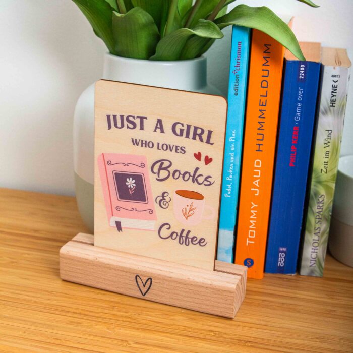 Eine rechteckige Holzpostkarte liegt flach auf einer hellbraunen Holzoberfläche mit deutlicher Maserung. Auf der Karte ist ein lila und rosa Grafikdesign mit dem englischen Spruch „JUST A GIRL WHO LOVES Books & Coffee“ aufgedruckt. Links neben dem Text „Books“ ist ein geschlossenes, rosa Buch mit einer kleinen Blume auf dem Cover und einem lila Lesebändchen abgebildet. Rechts neben dem Wort „Coffee“ ist eine weiße Kaffeetasse mit einer kleinen Pflanzen-Illustration zu sehen.