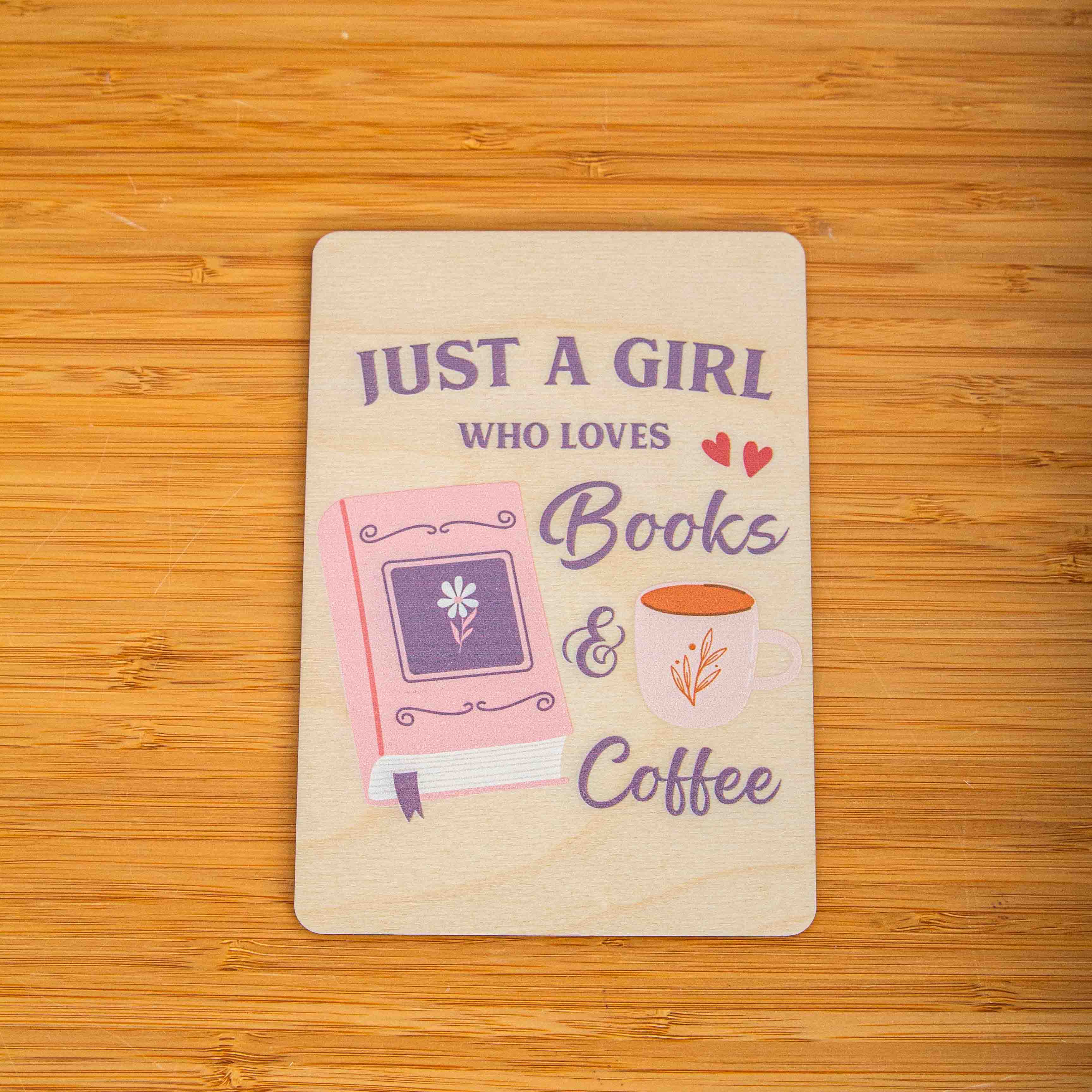 Postkarte Just a girl who loves books and coffee / Holzpostkarte Bücherliebhaber / Bookstagram / Postkarten mit Botschaft 3 Eine rechteckige Holzpostkarte liegt flach auf einer hellbraunen Holzoberfläche mit deutlicher Maserung. Auf der Karte ist ein lila und rosa Grafikdesign mit dem englischen Spruch „JUST A GIRL WHO LOVES Books & Coffee“ aufgedruckt. Links neben dem Text „Books“ ist ein geschlossenes, rosa Buch mit einer kleinen Blume auf dem Cover und einem lila Lesebändchen abgebildet. Rechts neben dem Wort „Coffee“ ist eine weiße Kaffeetasse mit einer kleinen Pflanzen-Illustration zu sehen.