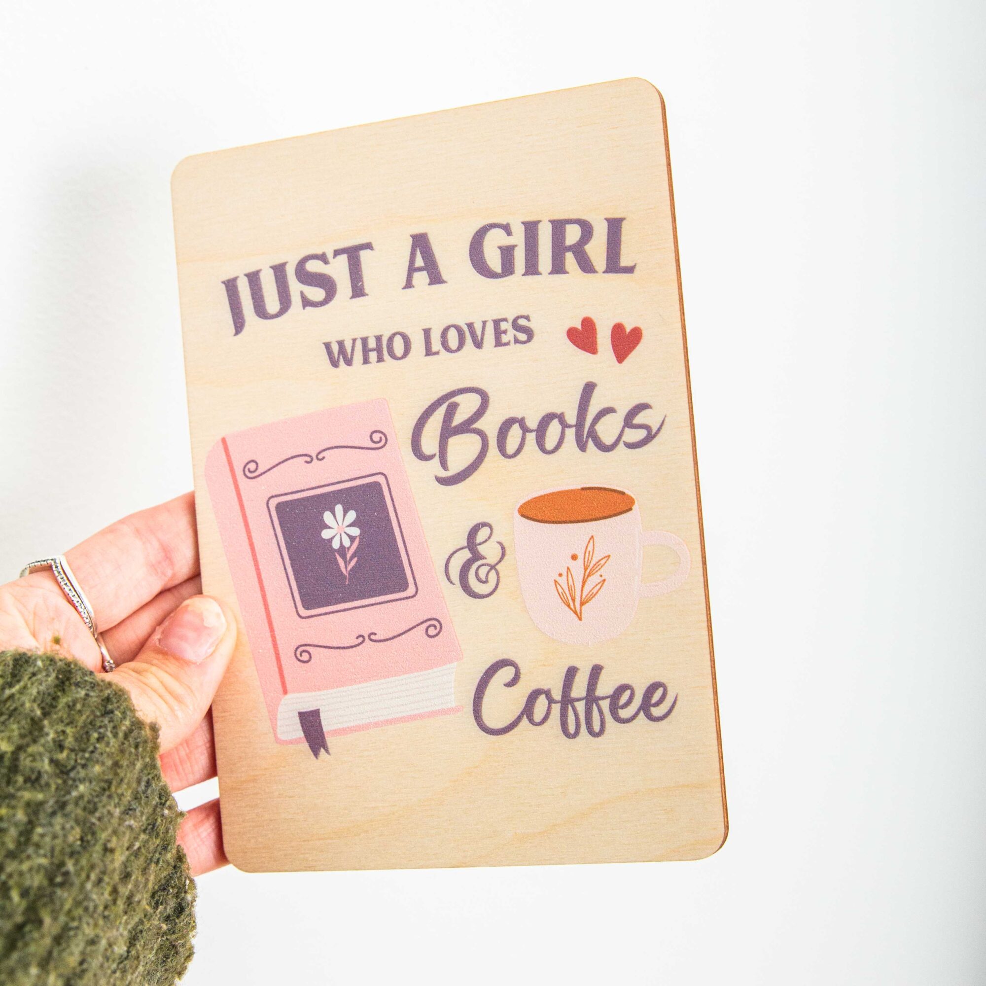Postkarte Just a girl who loves books and coffee / Holzpostkarte Bücherliebhaber / Bookstagram / Postkarten mit Botschaft 2 Eine rechteckige Holzpostkarte liegt flach auf einer hellbraunen Holzoberfläche mit deutlicher Maserung. Auf der Karte ist ein lila und rosa Grafikdesign mit dem englischen Spruch „JUST A GIRL WHO LOVES Books & Coffee“ aufgedruckt. Links neben dem Text „Books“ ist ein geschlossenes, rosa Buch mit einer kleinen Blume auf dem Cover und einem lila Lesebändchen abgebildet. Rechts neben dem Wort „Coffee“ ist eine weiße Kaffeetasse mit einer kleinen Pflanzen-Illustration zu sehen.