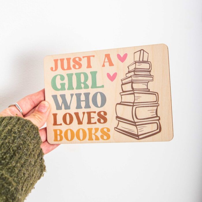 Eine rechteckige Holzpostkarte im Querformat liegt flach auf einer hellbraunen Holzoberfläche mit feiner Maserung. Auf der linken Seite der Karte steht in großen, farbigen Buchstaben im Retro-Stil der englische Text „JUST A GIRL WHO LOVES BOOKS“ untereinander geschrieben. Die Schriftfarben wechseln reihenweise von Lachsrosa über Mintgrün, Graublau und Braun bis hin zu Gelb. Rechts daneben ist eine schlichte, braune Liniengrafik eines hohen Bücherstapels abgebildet.
