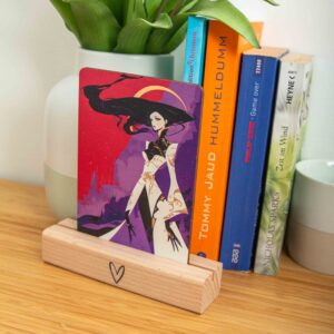 Geschenke und Deko 17 Eine rechteckige Holzpostkarte im Hochformat liegt flach auf einer hellbraunen Holzoberfläche. Die Karte zeigt eine vollflächige, dramatische Fantasy-Illustration im modernen, kantigen Graphic-Novel-Stil. Im Zentrum steht eine elegante weibliche Figur mit extrem langem, fließendem schwarzen Haar und auffälligem roten Make-up. Sie trägt ein stilisiertes weißes Gewand mit goldenen Ornamenten und weiten Ärmeln, kombiniert mit schwarzen Elementen und langen, spitzen Fingern.