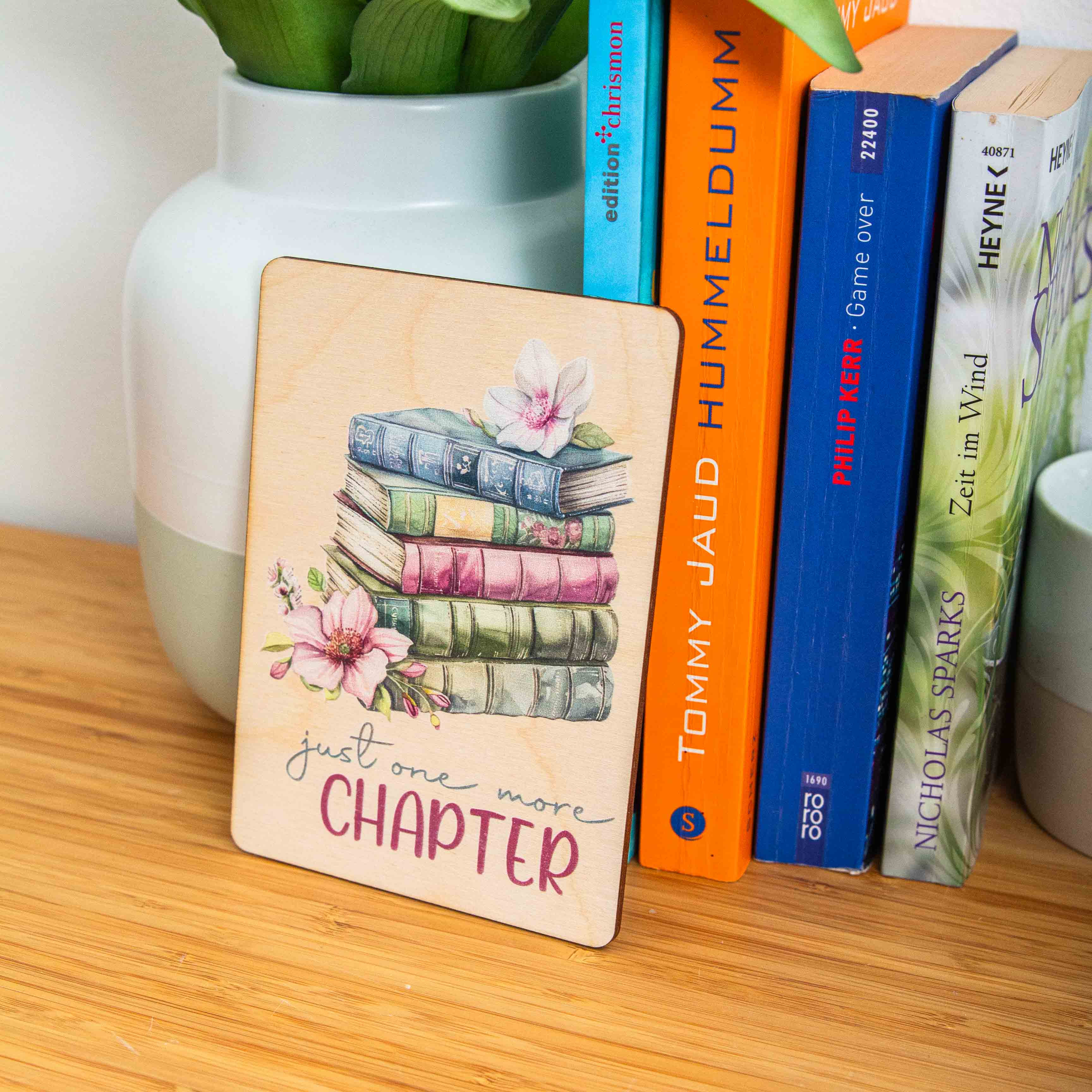 Postkarte Just one more chapter / Holzpostkarte Bücherliebhaber / Bookstagram / Postkarten mit Botschaft 4 Eine Hand in einem olivgrünen Strickpullover hält eine hochkant ausgerichtete Holzpostkarte vor einem weißen Hintergrund. Auf dem Holz befindet sich eine zarte Illustration im Vintage-Stil, die einen Stapel von fünf alten Büchern in gedeckten Blau-, Grün- und Rosatönen zeigt. Auf dem obersten Buch liegt eine helle Blüte, und links neben dem Stapel ist eine weitere, rosa Magnolienblüte zu sehen. Darunter steht in geschwungener Schrift und Druckbuchstaben der englische Spruch:just one more CHAPTER