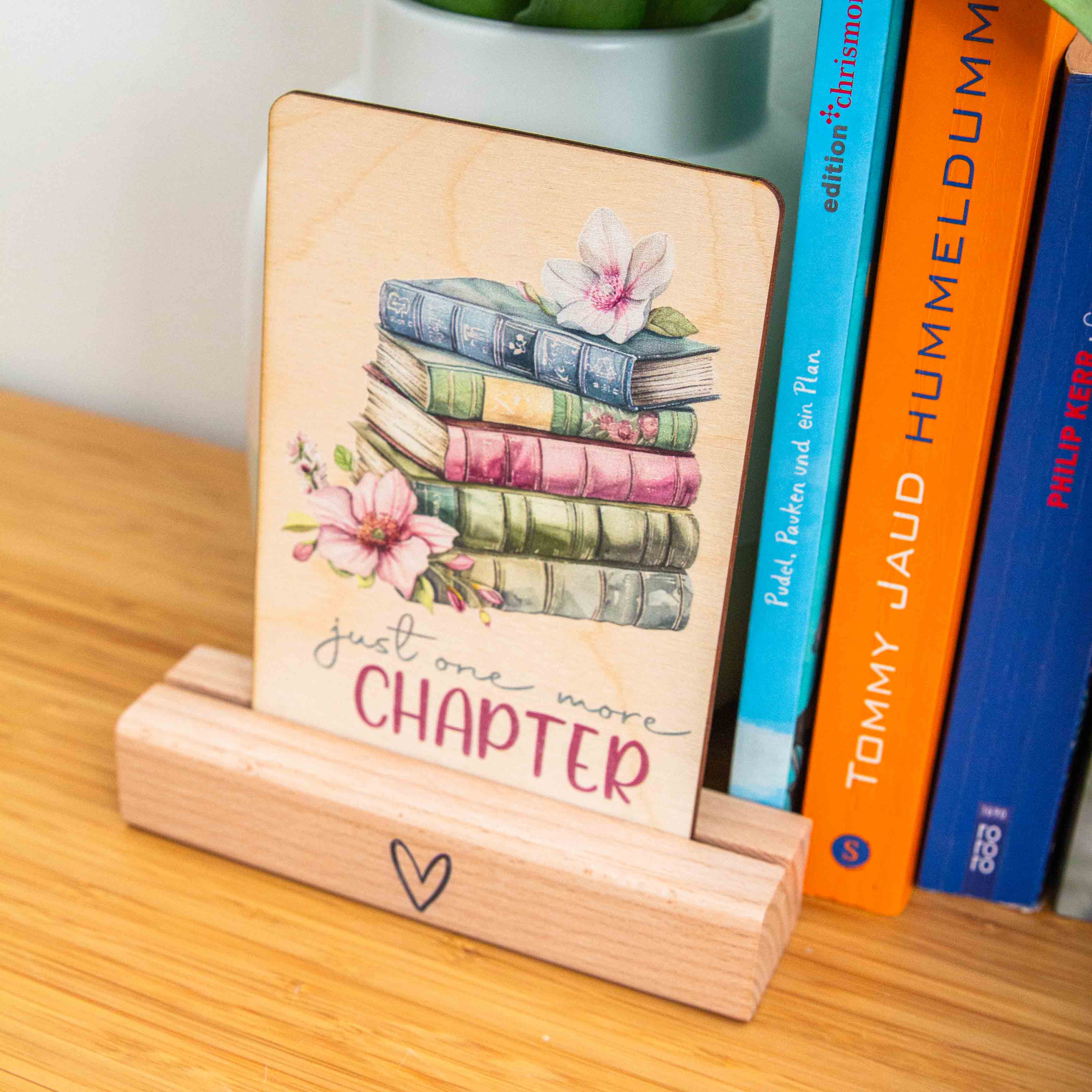 Postkarte Just one more chapter / Holzpostkarte Bücherliebhaber / Bookstagram / Postkarten mit Botschaft 1 Eine Hand in einem olivgrünen Strickpullover hält eine hochkant ausgerichtete Holzpostkarte vor einem weißen Hintergrund. Auf dem Holz befindet sich eine zarte Illustration im Vintage-Stil, die einen Stapel von fünf alten Büchern in gedeckten Blau-, Grün- und Rosatönen zeigt. Auf dem obersten Buch liegt eine helle Blüte, und links neben dem Stapel ist eine weitere, rosa Magnolienblüte zu sehen. Darunter steht in geschwungener Schrift und Druckbuchstaben der englische Spruch:just one more CHAPTER