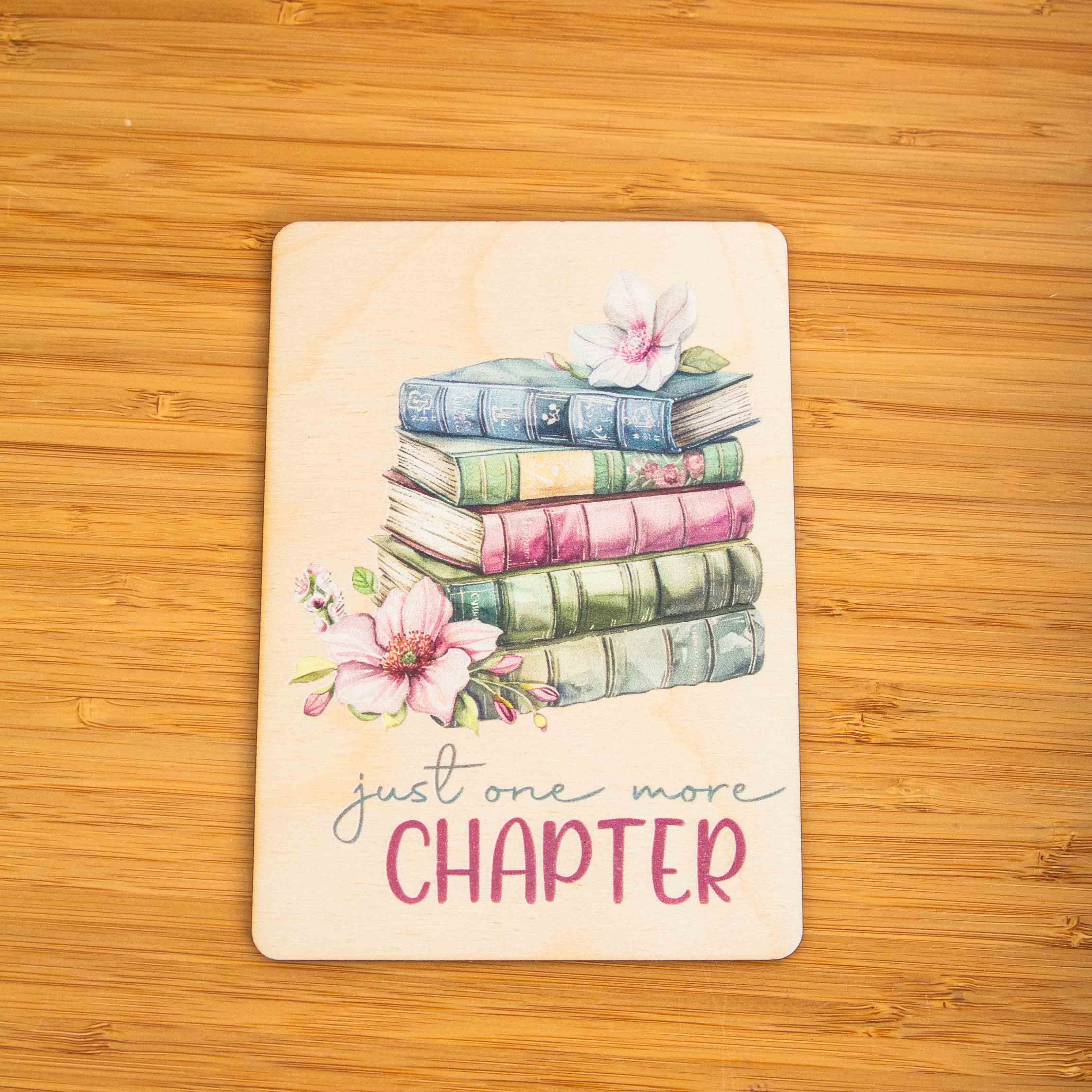 Postkarte Just one more chapter / Holzpostkarte Bücherliebhaber / Bookstagram / Postkarten mit Botschaft 3 Eine Hand in einem olivgrünen Strickpullover hält eine hochkant ausgerichtete Holzpostkarte vor einem weißen Hintergrund. Auf dem Holz befindet sich eine zarte Illustration im Vintage-Stil, die einen Stapel von fünf alten Büchern in gedeckten Blau-, Grün- und Rosatönen zeigt. Auf dem obersten Buch liegt eine helle Blüte, und links neben dem Stapel ist eine weitere, rosa Magnolienblüte zu sehen. Darunter steht in geschwungener Schrift und Druckbuchstaben der englische Spruch:just one more CHAPTER