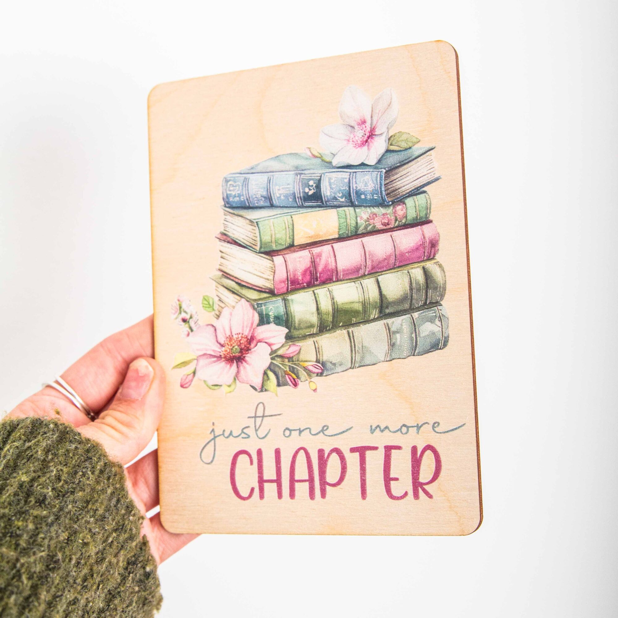 Postkarte Just one more chapter / Holzpostkarte Bücherliebhaber / Bookstagram / Postkarten mit Botschaft 2 Eine Hand in einem olivgrünen Strickpullover hält eine hochkant ausgerichtete Holzpostkarte vor einem weißen Hintergrund. Auf dem Holz befindet sich eine zarte Illustration im Vintage-Stil, die einen Stapel von fünf alten Büchern in gedeckten Blau-, Grün- und Rosatönen zeigt. Auf dem obersten Buch liegt eine helle Blüte, und links neben dem Stapel ist eine weitere, rosa Magnolienblüte zu sehen. Darunter steht in geschwungener Schrift und Druckbuchstaben der englische Spruch:just one more CHAPTER